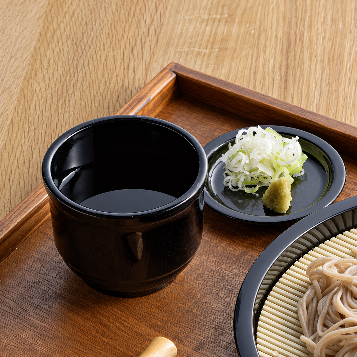 Soba dipping cup filled with dipping sauce, condiment dish with spring onion and wasabi beside a zaru soba plate